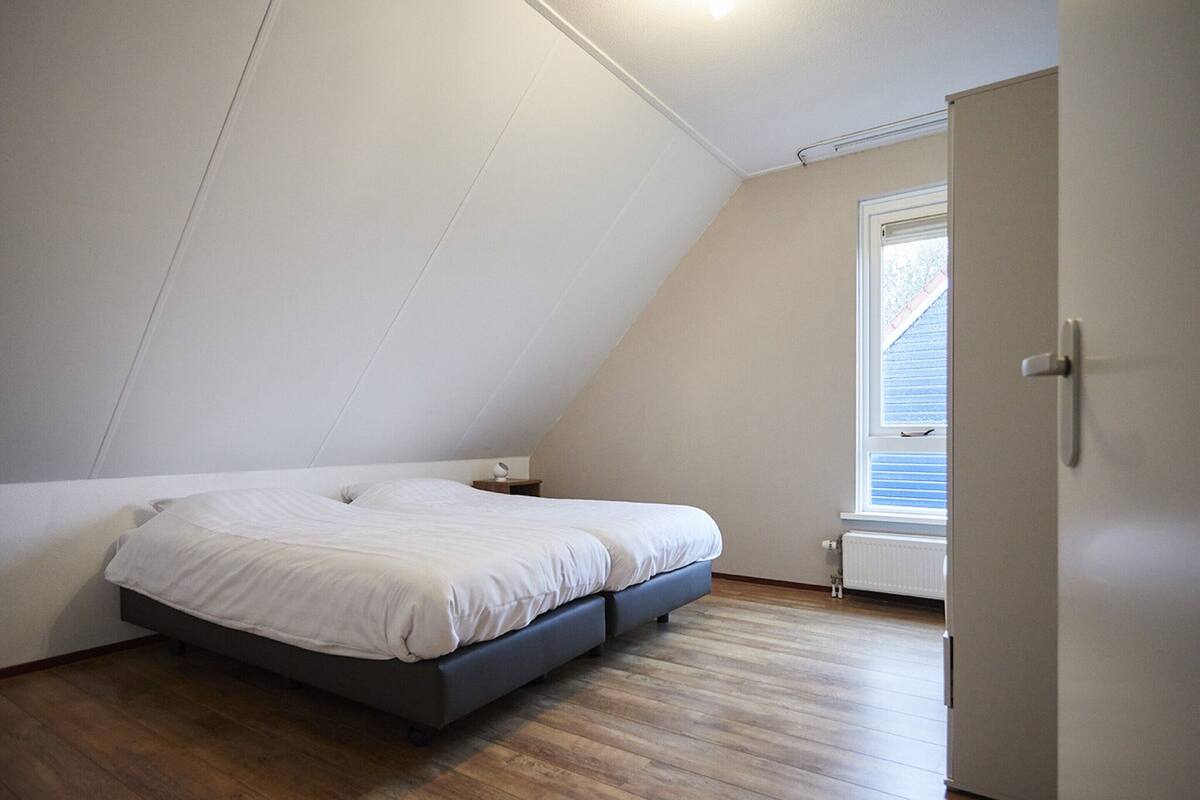 Une chambre avec un plafond en pente, un lit avec une literie blanche, une table de chevet et une fenêtre avec des stores.