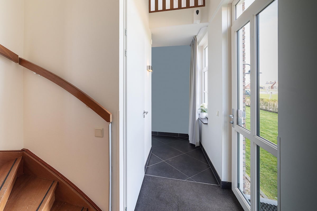 Un couloir avec un escalier en bois, une porte donnant sur une pièce et de grandes fenêtres offrant une vue sur l’extérieur.