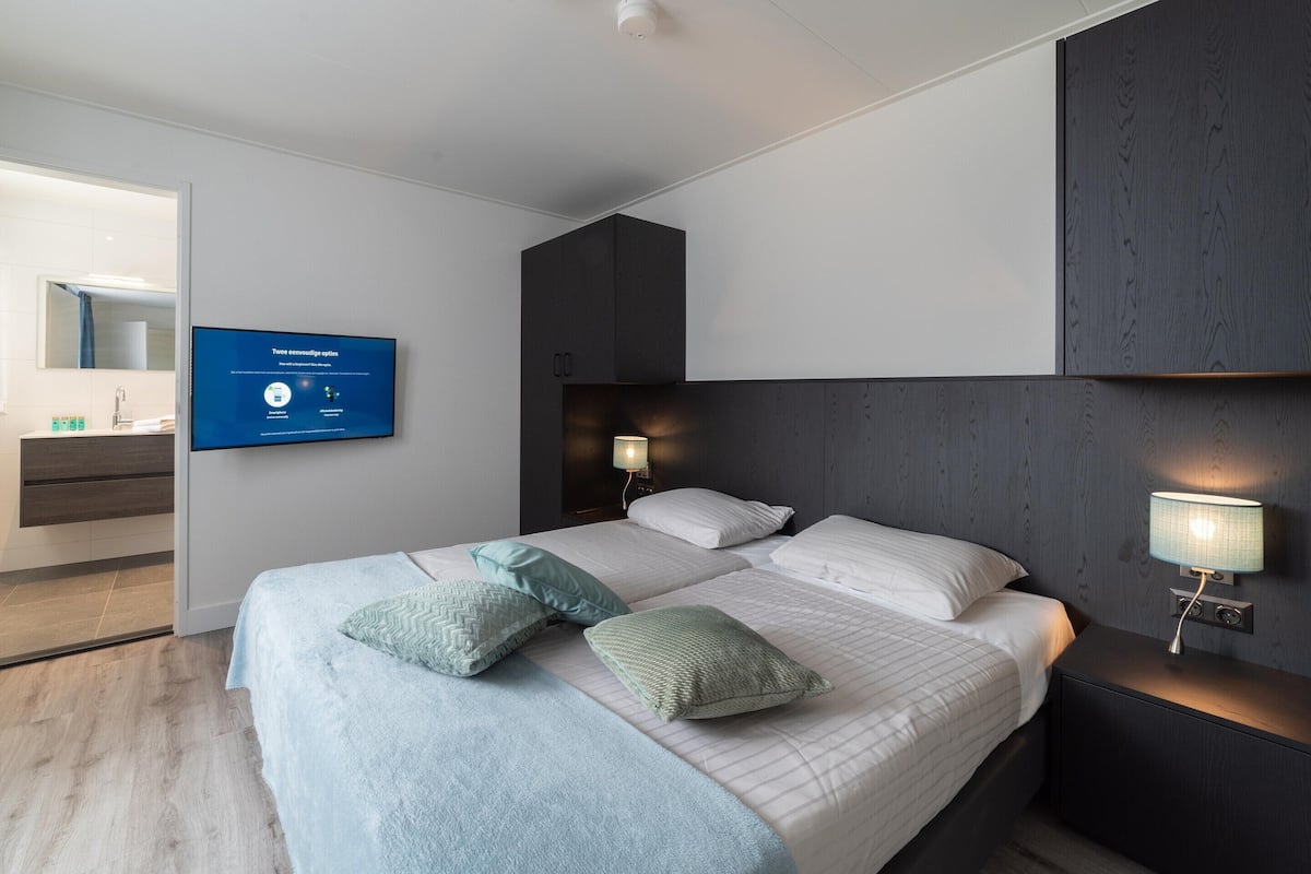 Une chambre d’hôtel moderne avec un grand lit, un téléviseur à écran plat fixé au mur, une table de chevet avec une lampe et une tête de lit en bois.