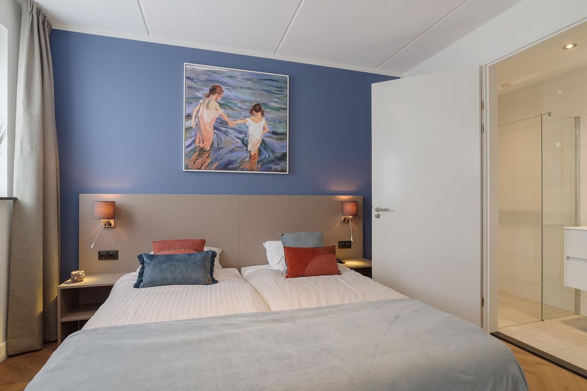Une chambre d’hôtel avec deux lits, un tableau représentant deux personnages accroché au mur, des lampes de chevet et une porte de douche en verre.