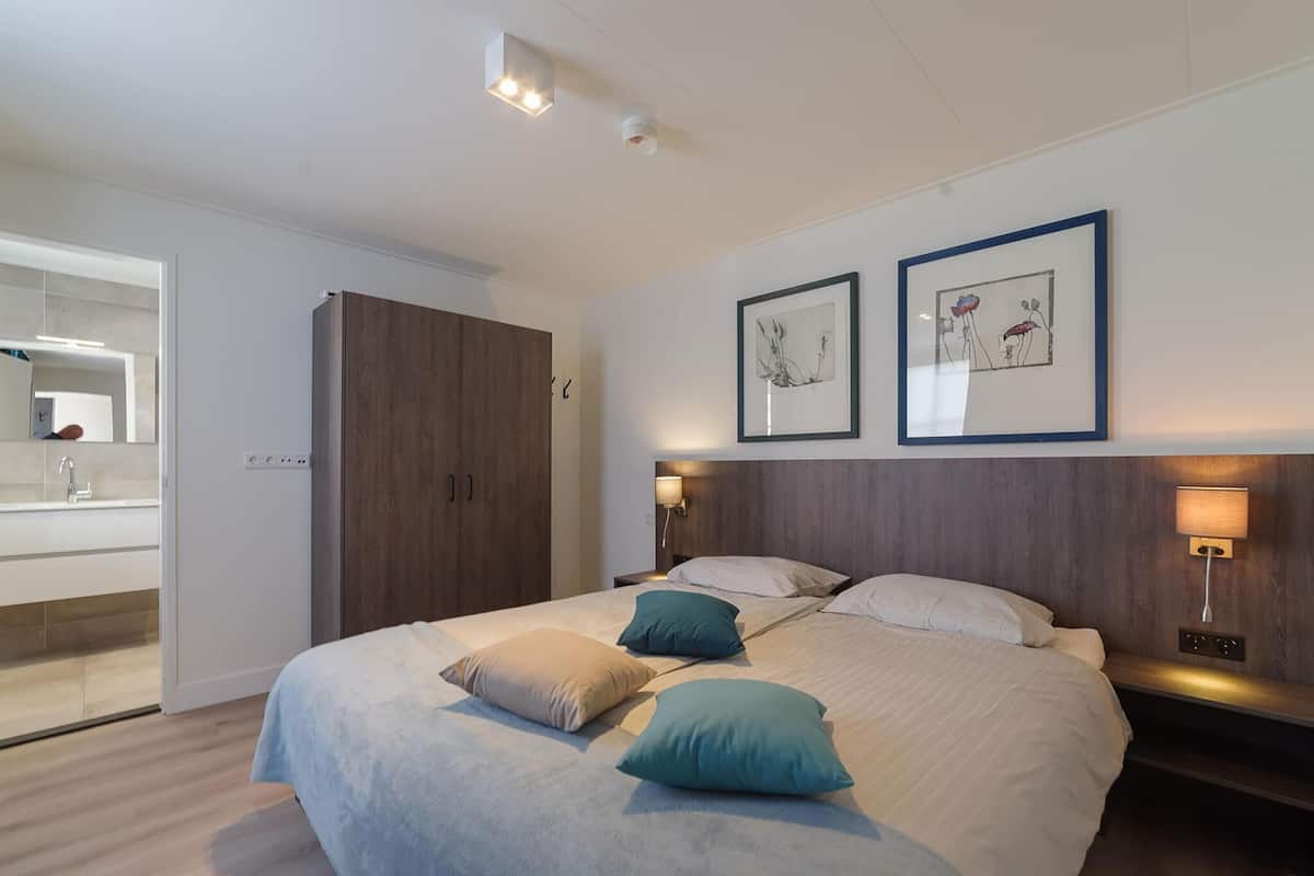 Une chambre moderne avec un grand lit, des lampes de chevet, une armoire et une salle de bain visible à travers une porte ouverte.