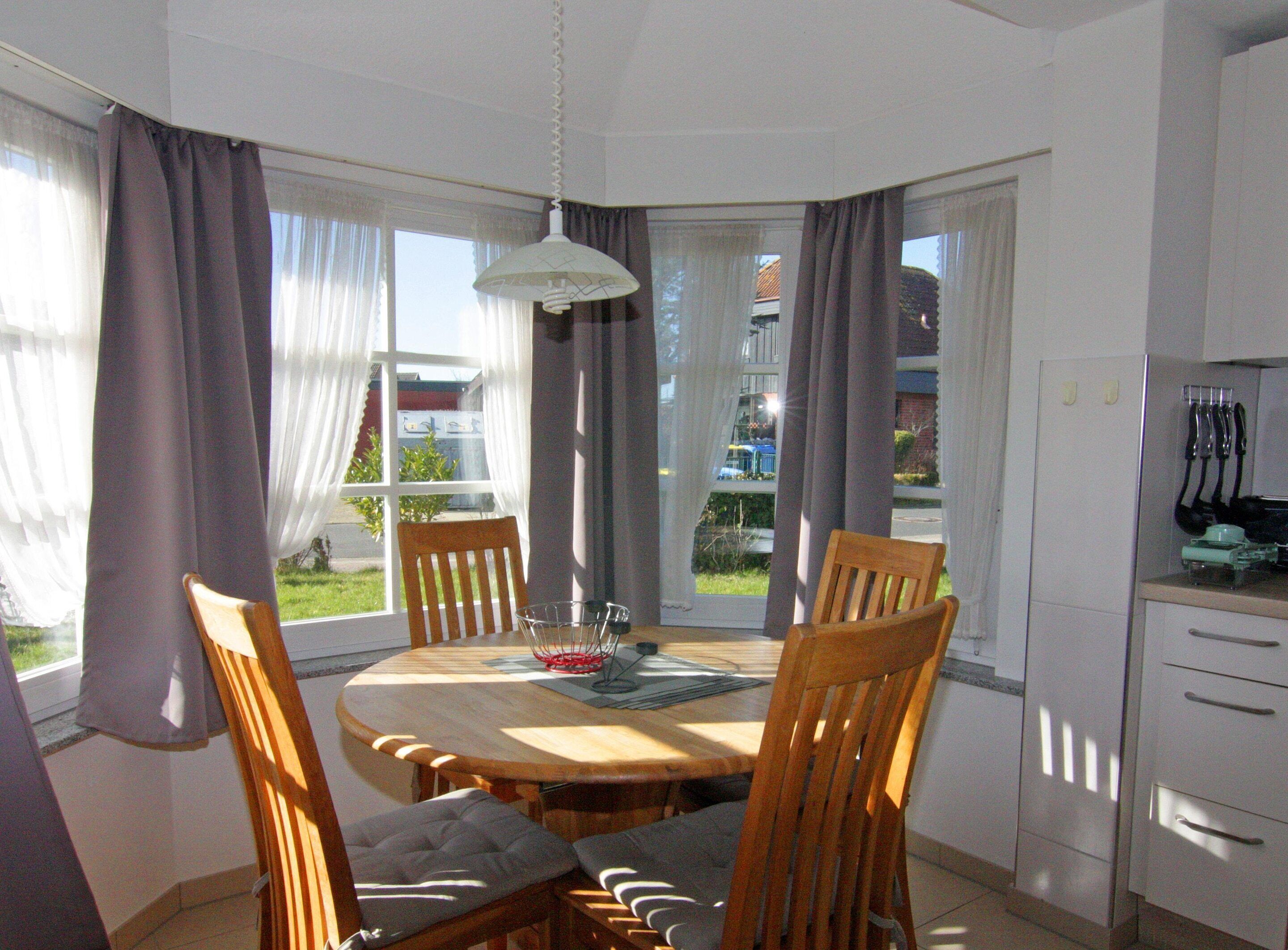 Ein Essbereich mit einem runden Tisch und Stühlen, einer hängenden Leuchte und Blick durch große, mit Vorhängen drapierten Fenster nach draußen.