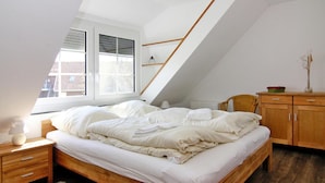 Une chambre avec un plafond en pente, un lit en bois, une table de chevet, une chaise, une commode et une fenêtre avec des stores.