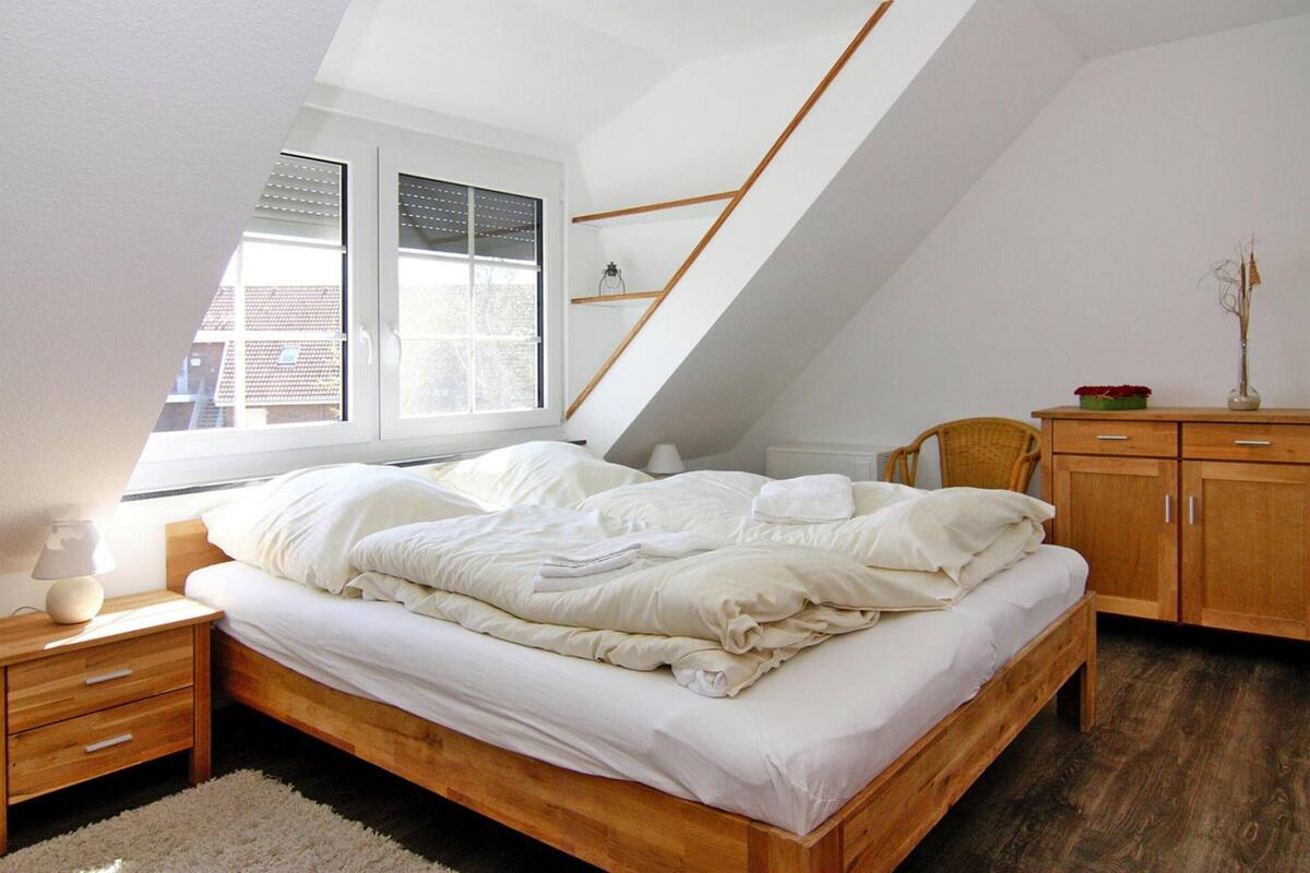 A bedroom with a sloped ceiling, a bed with white bedding, a nightstand with a lamp, a wooden dresser, and a window with blinds.