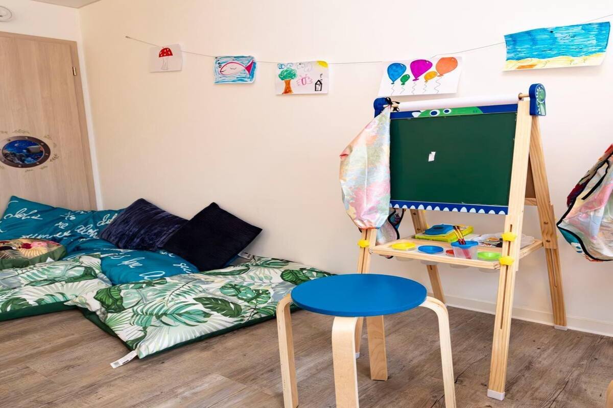 Una habitación acogedora con suelo de madera, un caballete verde con un lienzo en blanco, una silla azul y una cama con ropa de cama azul y verde.