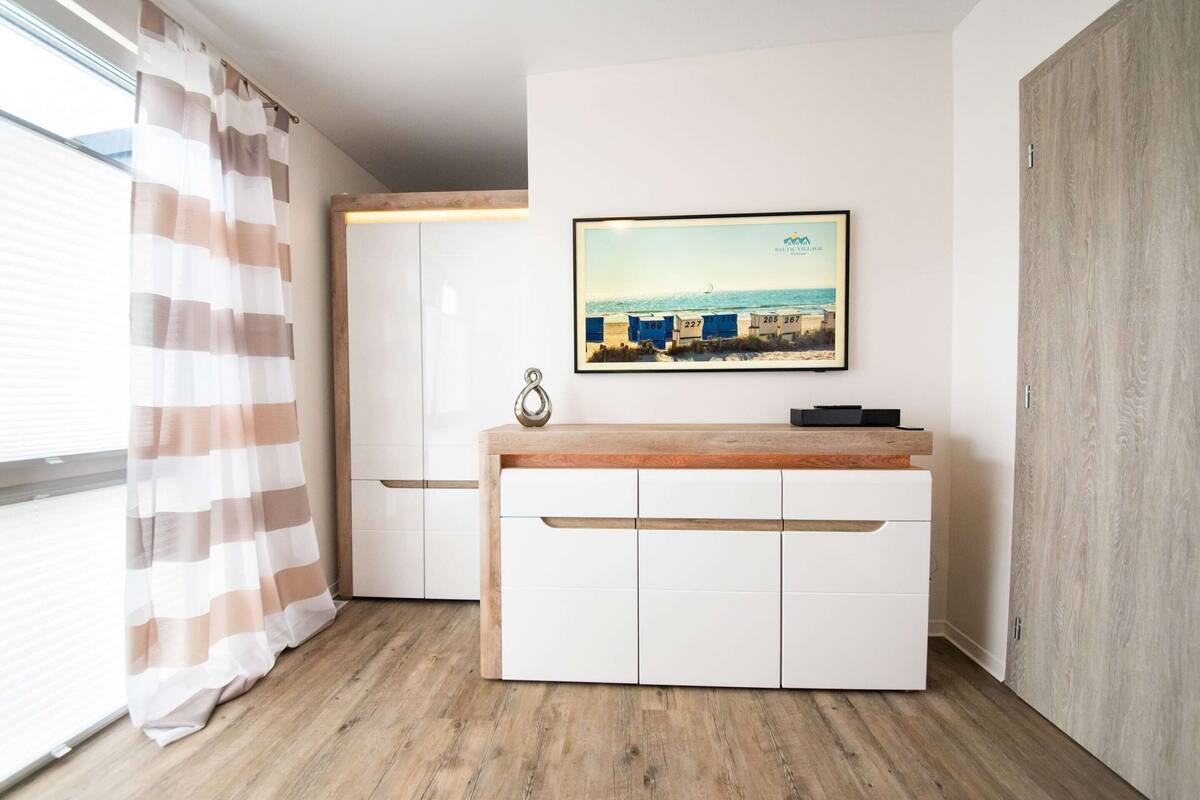 Una habitación moderna con piso de madera, muebles de color blanco y un cuadro en la pared, además de una cortina a rayas.