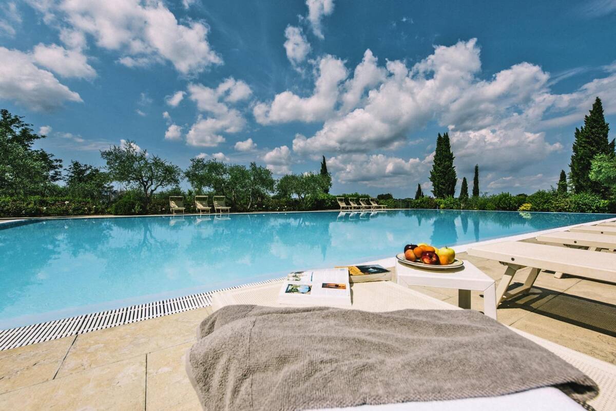 A poolside area with lounge chairs, a table with fruits, and a view of the pool and trees under a partly cloudy sky.