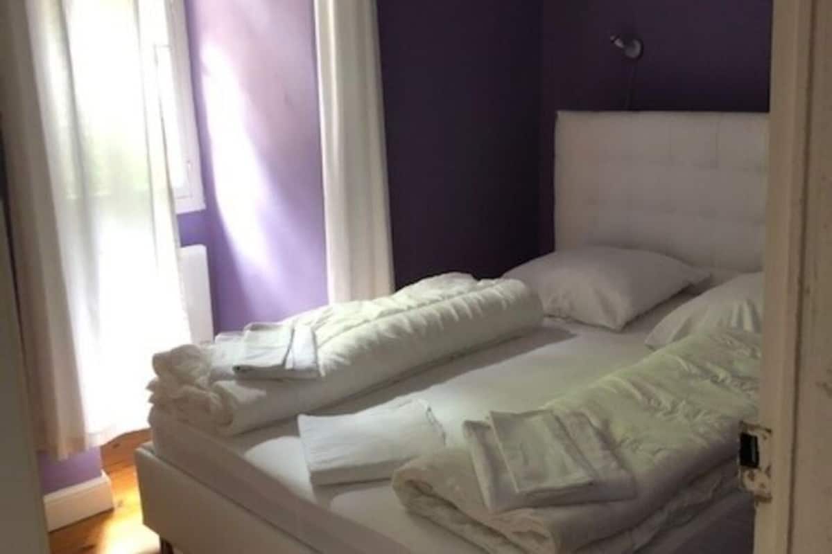 Een tweepersoonsbed met wit beddengoed in een kamer met paarse wanden en een raam met gordijnen.