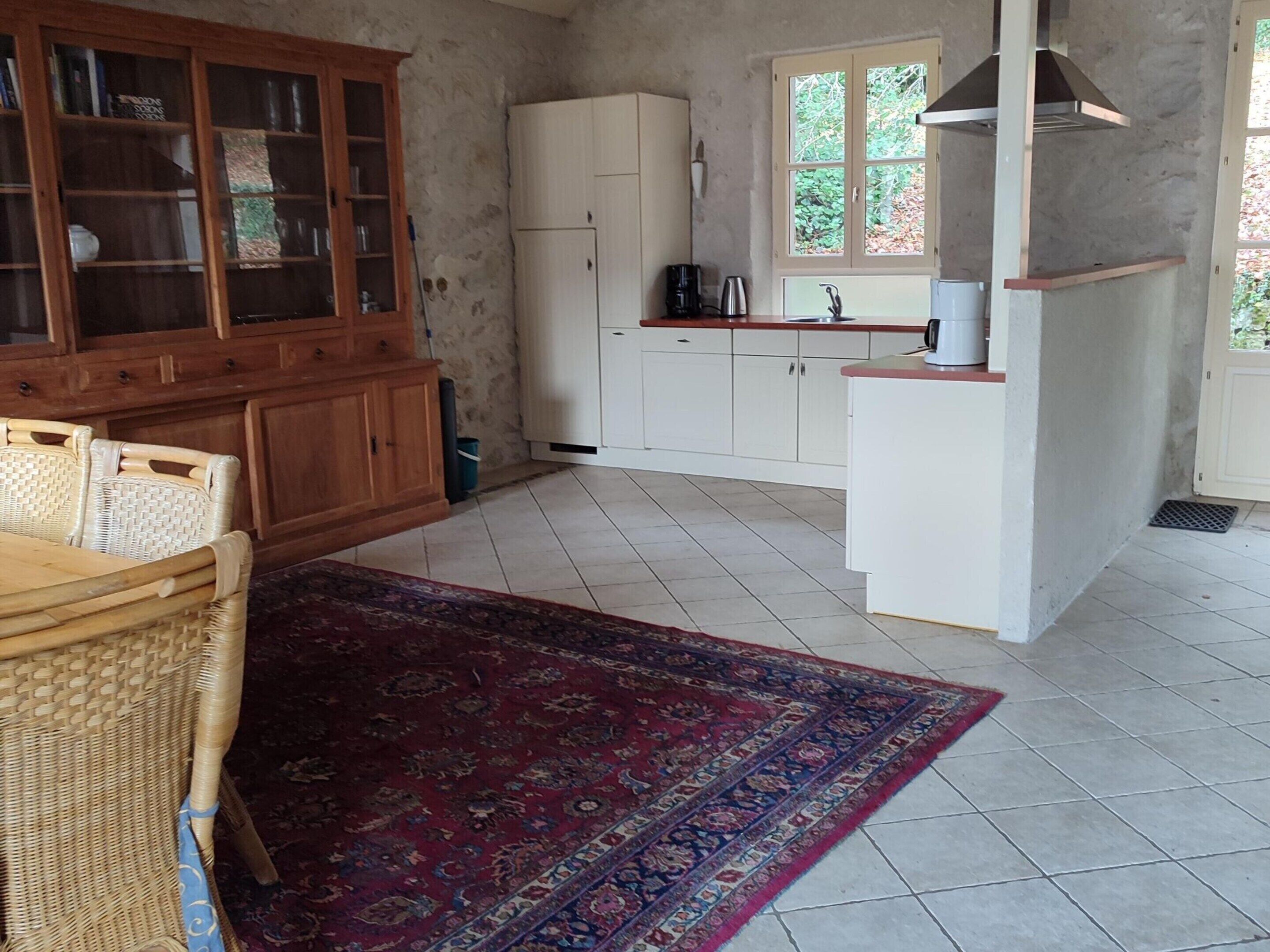 Una cucina con mobili bianchi, un tavolo da pranzo in legno con sedie, un tappeto rosso decorato e una finestra vista verde.