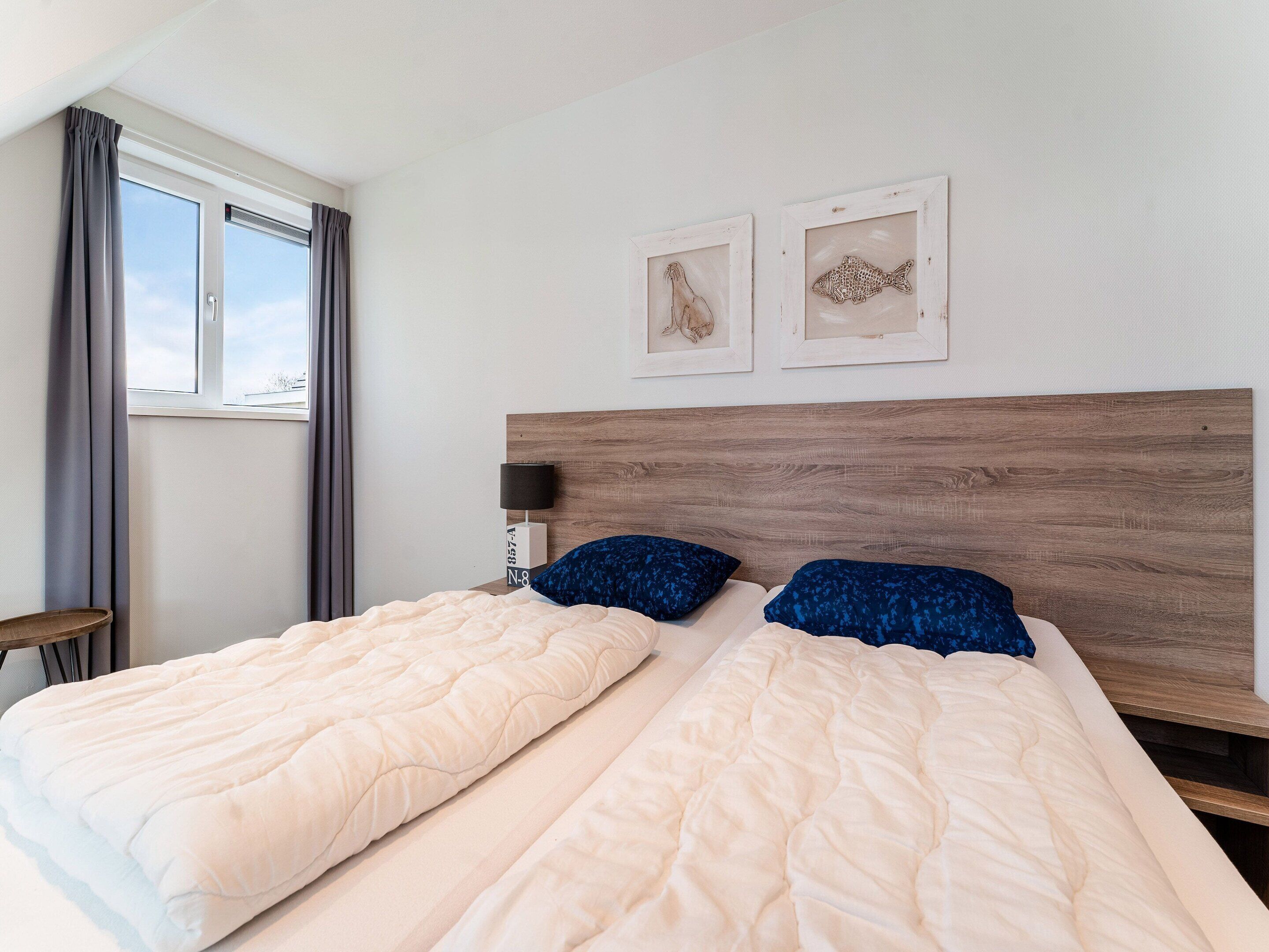 A double bed with a wooden headboard, two pillows, a bedside table with a lamp, and two framed artworks on the wall.