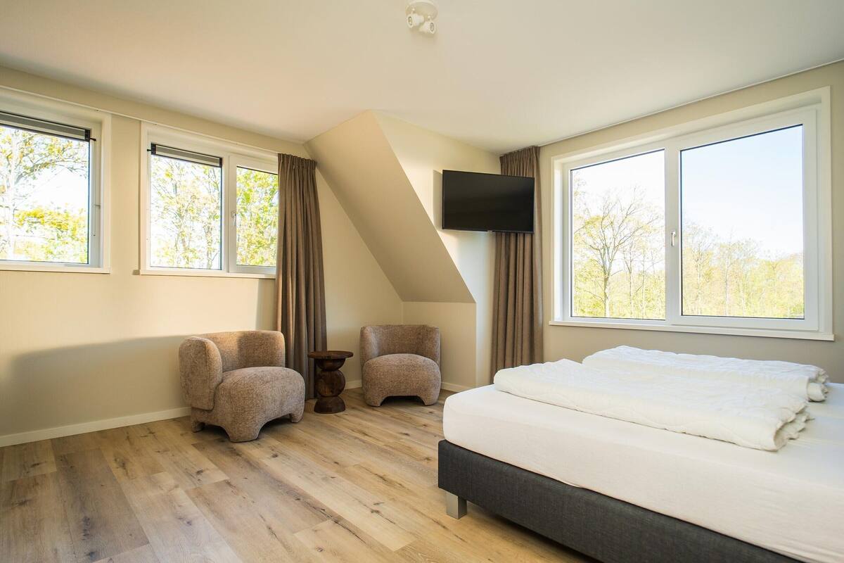 Een slaapkamer met een bed, twee fauteuils, een tafeltje, een televisie en grote ramen.