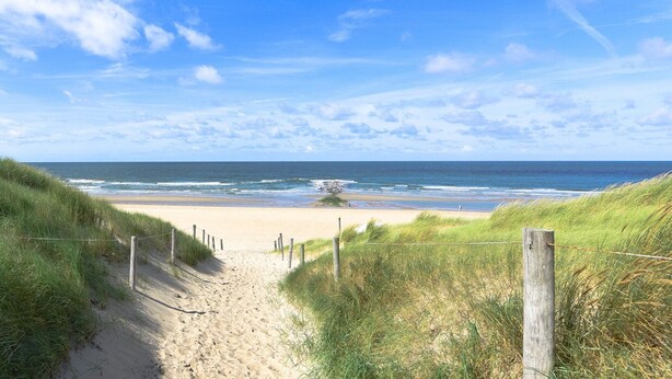 Un sentier de sable qui longe la plage et mène à l’océan, bordé de dunes herbeuses et d’une clôture en bois.