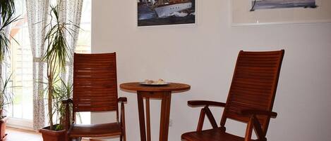 A room with two wooden chairs, a small round table, and a potted plant. There are two framed pictures on the wall.
