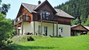 A two-story wooden house with a balcony, surrounded by a lush green landscape and a forested hill in the background. - Landhaus Amadeus, Radstadt (Radstadt)