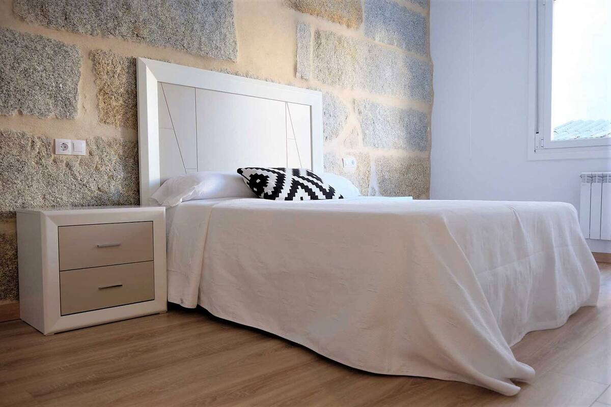 Une chambre avec un mur en pierre, un lit blanc avec un oreiller à motifs et une table de chevet avec des tiroirs.