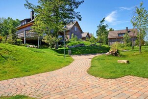 A brick pathway leading to a row of rustic-style houses with a grassy hill and trees in the background.