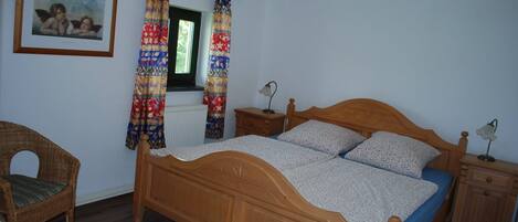 A bedroom with a wooden bed, a wicker chair, a nightstand, a bedside table, a lamp, and a window with curtains.