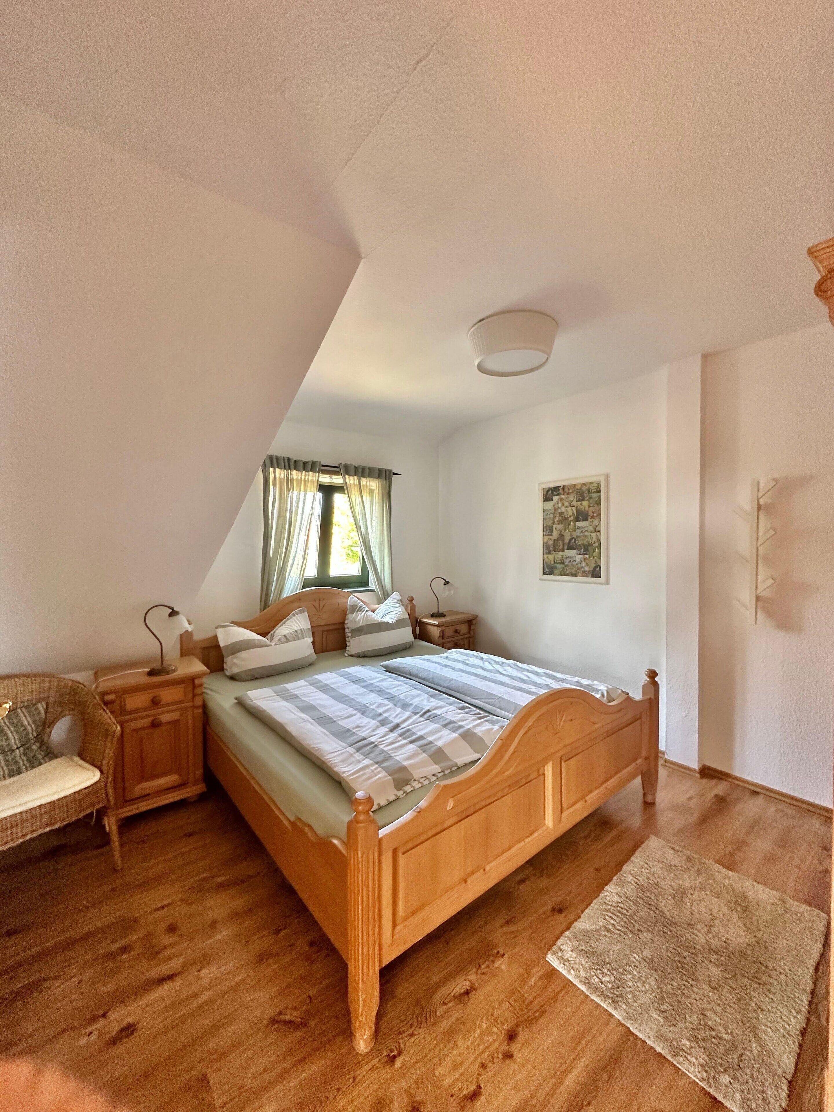 Un dormitorio con una cama de madera, una mesita de noche, una silla, un ventanal con cortinas y un cuadro en la pared.