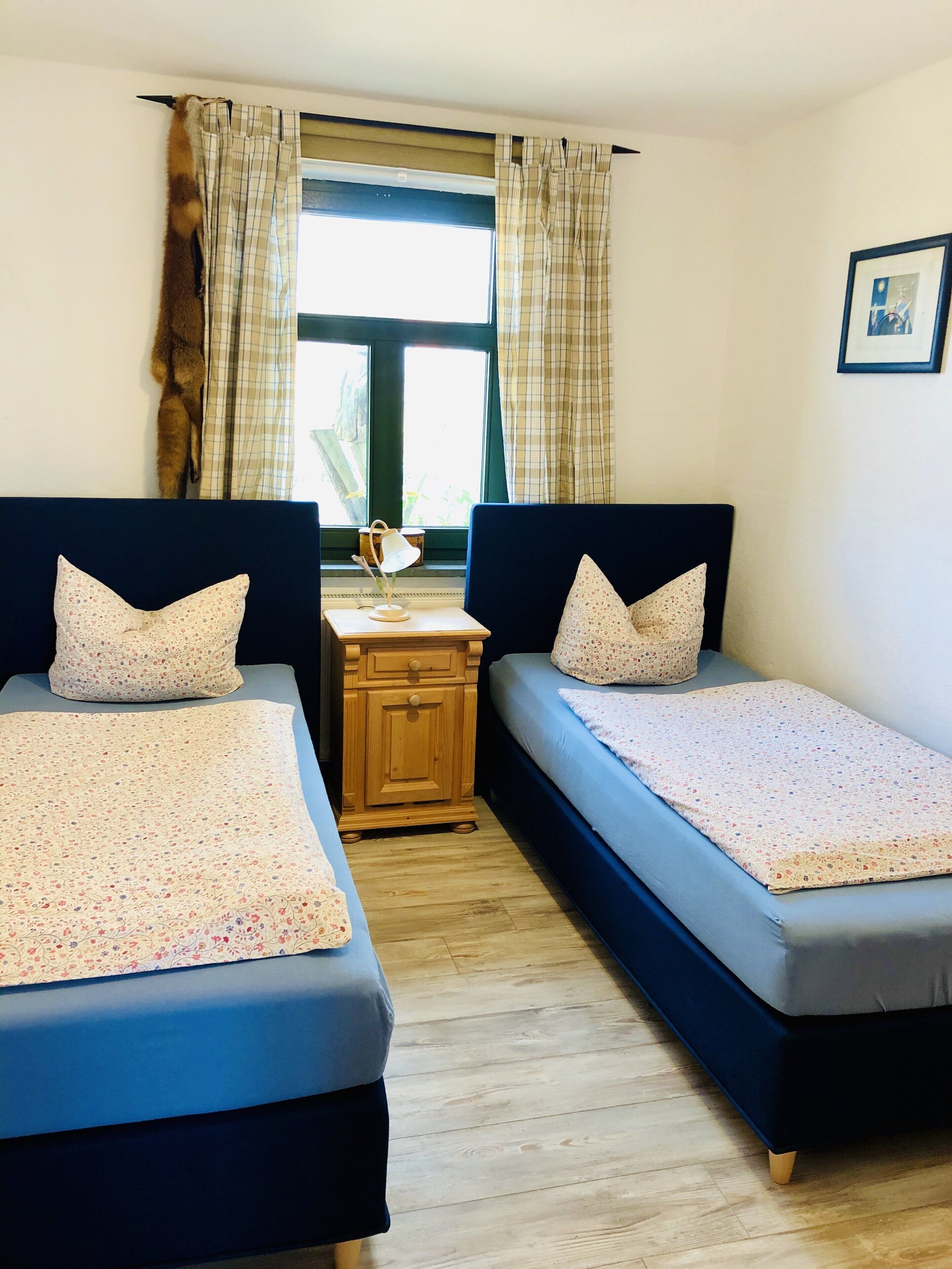 A room with two single beds, a wooden nightstand, a lamp, and a window with curtains.