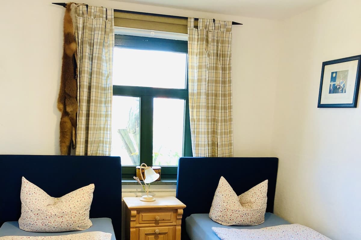 A room with two single beds, a wooden nightstand, a small table with a lamp, and a window with curtains.