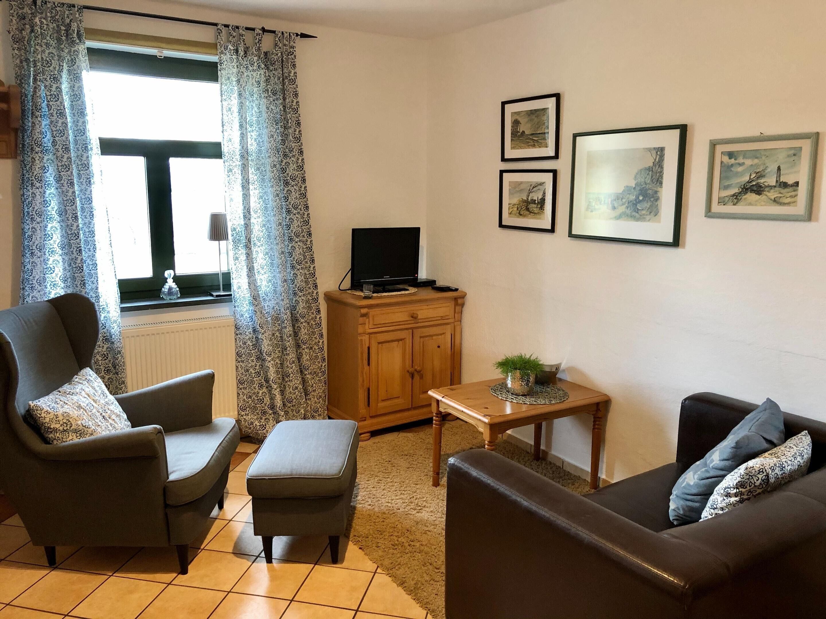 Een woonkamer met een bank, fauteuil, voetenbankje, houten kast, televisie en ingelijste foto's aan de muur.