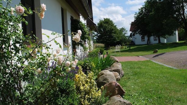 A building with a white facade and dark trim, surrounded by a well-maintained garden with various plants and flowers. - Grosses Landhaus mit Swimmingpool in Dargun (Dargun)