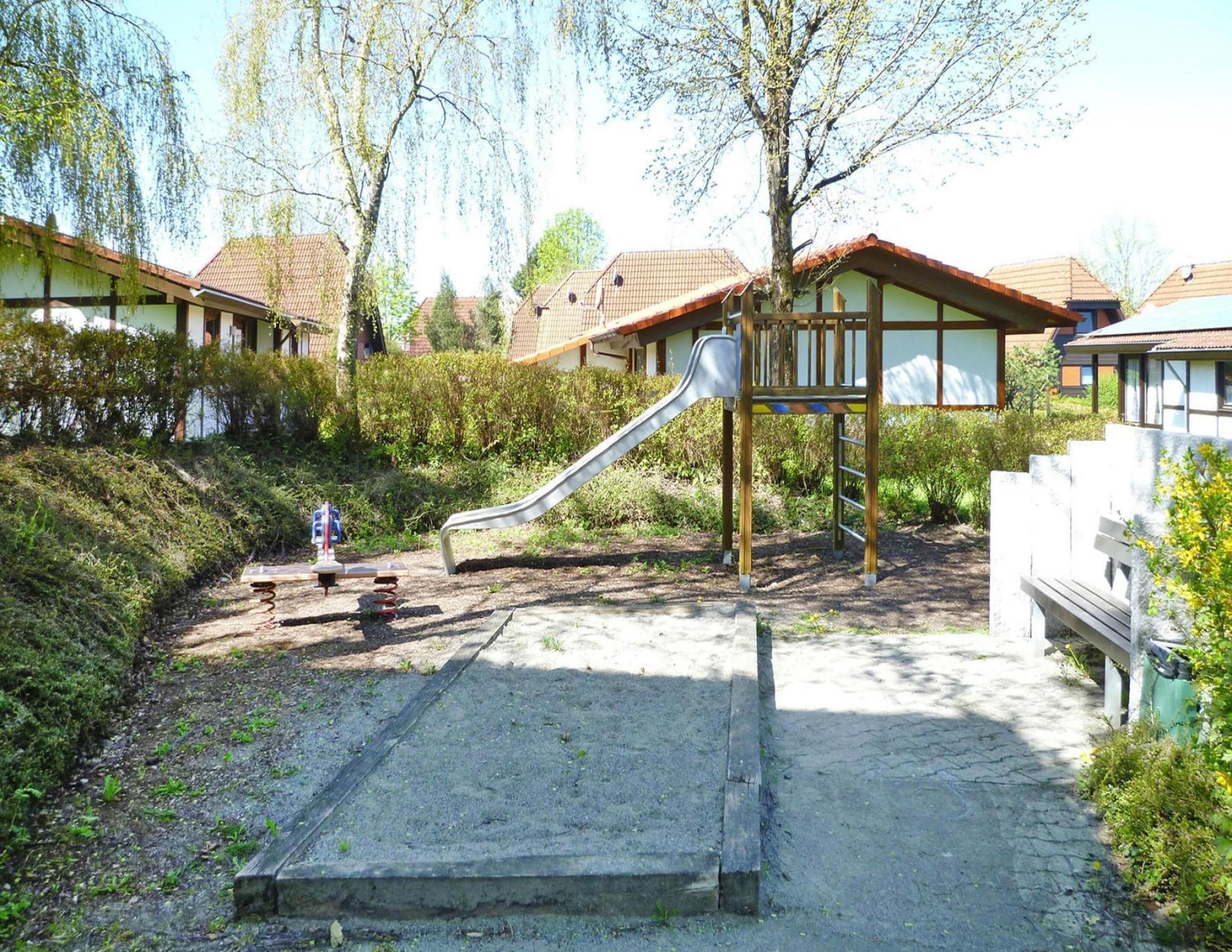 Un parque infantil con un tobogán y un banco pequeño, rodeado de vegetación y edificios residenciales.