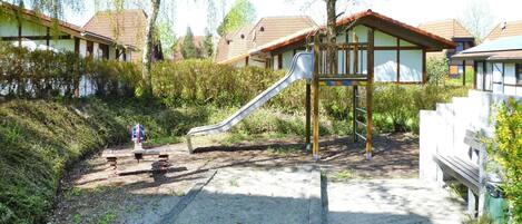 Un parque infantil con un tobogán y un banco pequeño, rodeado de vegetación y edificios residenciales.