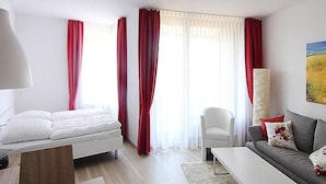 Ett modernt hotellrum med en säng, en soffa, ett soffbord och en stol. Det finns gardiner vid fönstren och en tavla på väggen.