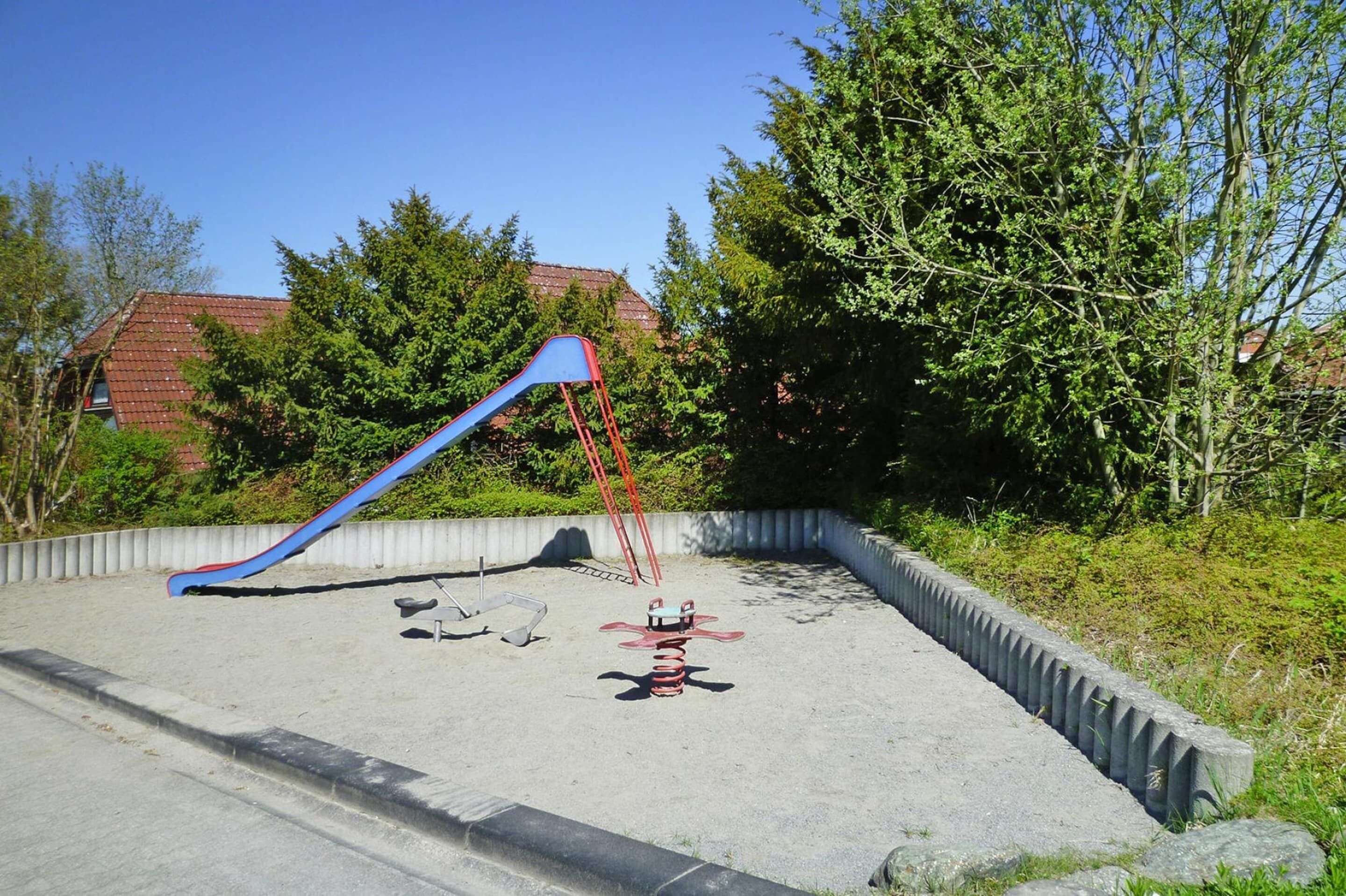 A playground with a blue slide and red climbing structure, surrounded by trees and a building in the background.