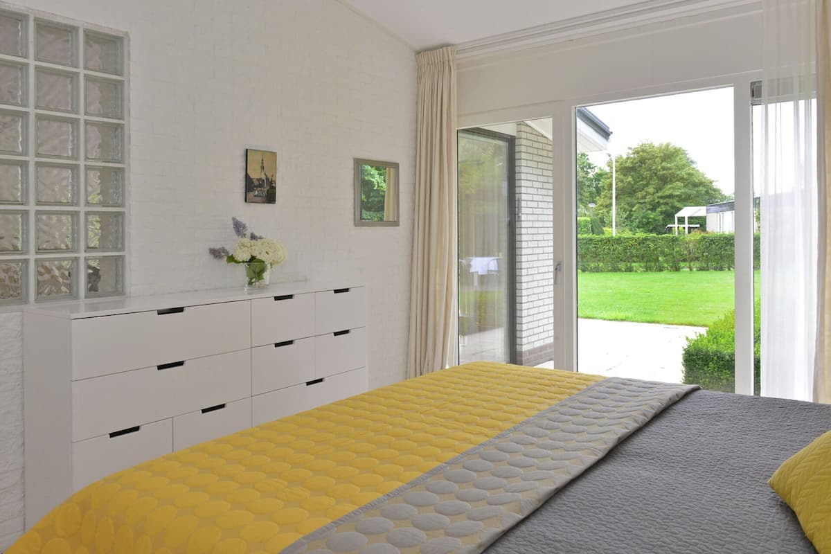 Un dormitorio con cama, cómoda, ventana con vista a follaje y puerta que da al exterior.