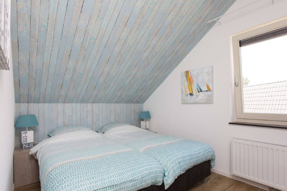 A bedroom with a sloped ceiling, a bed with a patterned bedspread, two bedside lamps, a radiator, and a window with a view of sailboats.