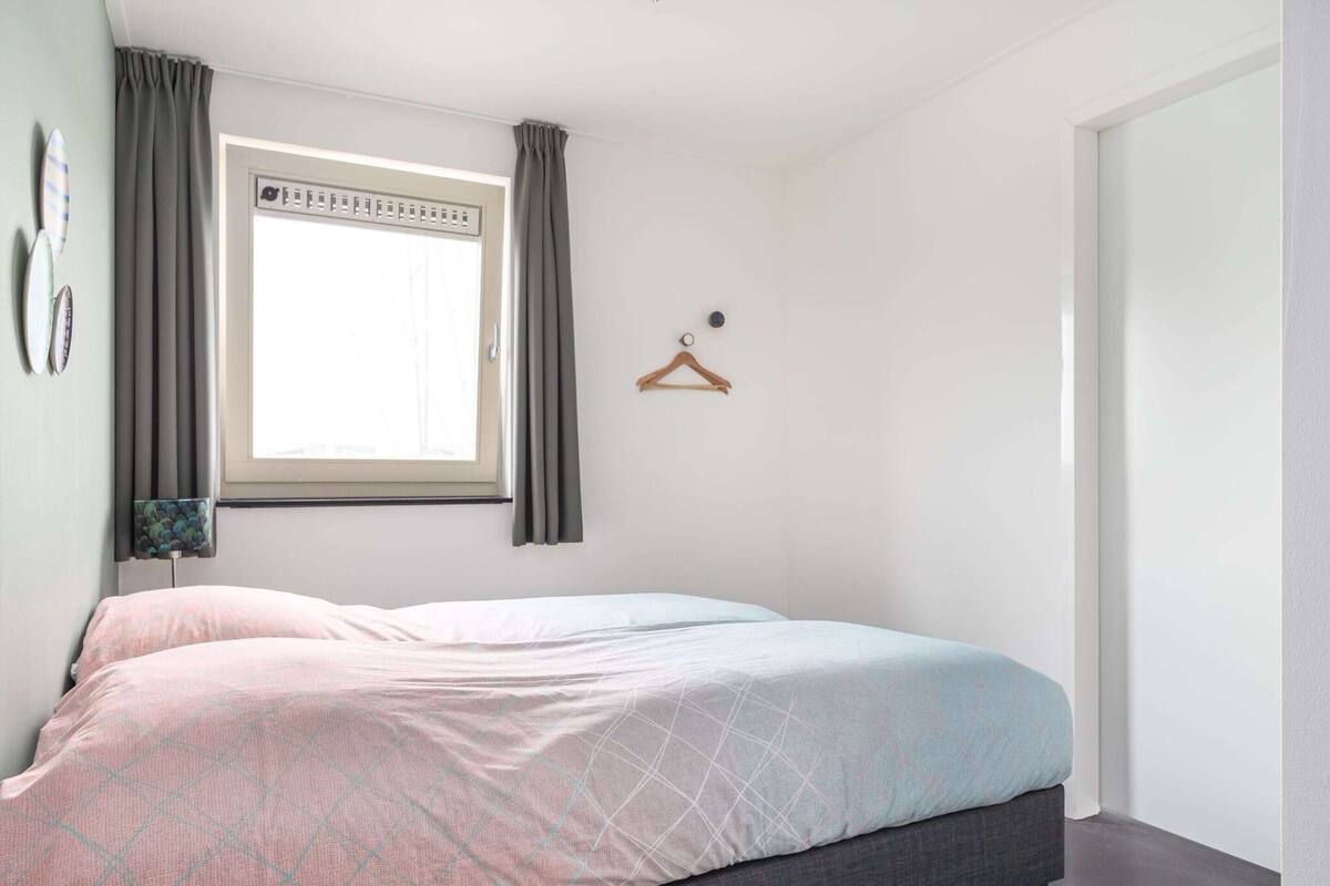 A bedroom with a bed, a window with curtains, a bedside table with a lamp, and a door.
