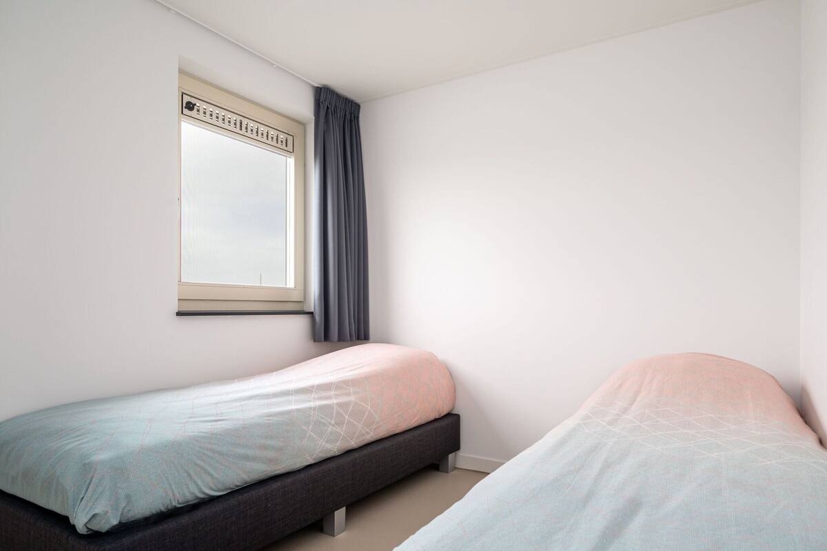 A small, single-room bedroom with two beds, a window with a curtain, and a plain white wall.