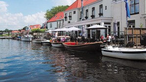 A waterfront area with boats docked along the water, a row of buildings with red roofs, and outdoor seating areas with umbrellas. - Bungalow With an Outdoor Fireplace by Water (Akkrum)