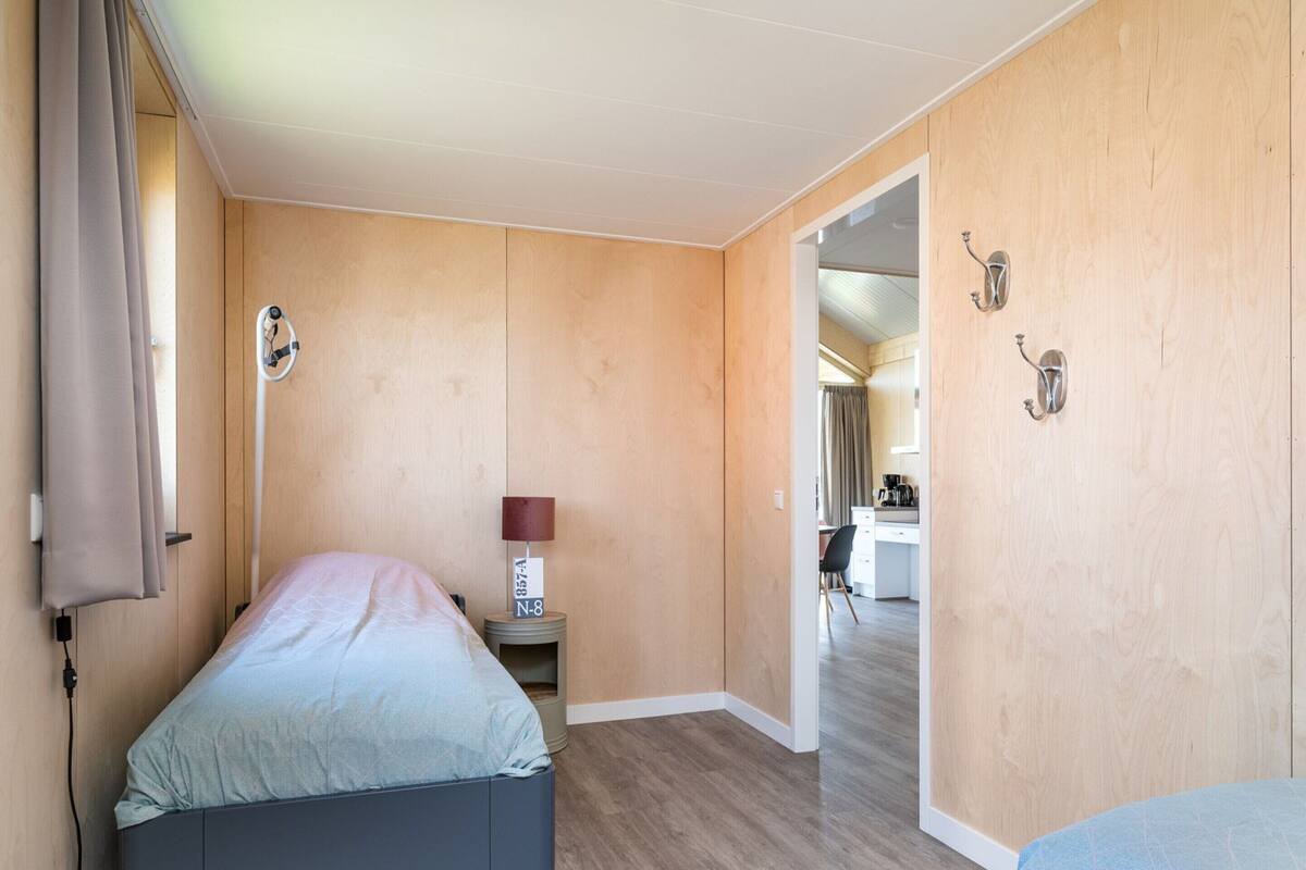 A bedroom with a bed, bedside table, lamp, and a view into a kitchen area.