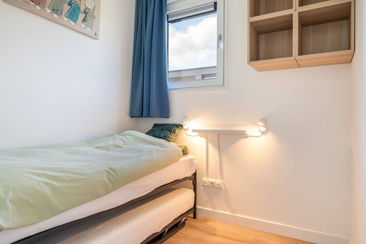 A single bed with a headboard, a bedside table with a lamp, a window with curtains, and a wall-mounted shelf.