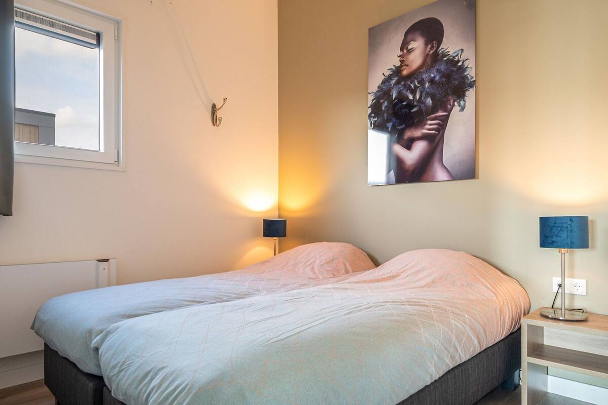 A bedroom with a bed, a nightstand, a wall-mounted lamp, a window, and a painting.