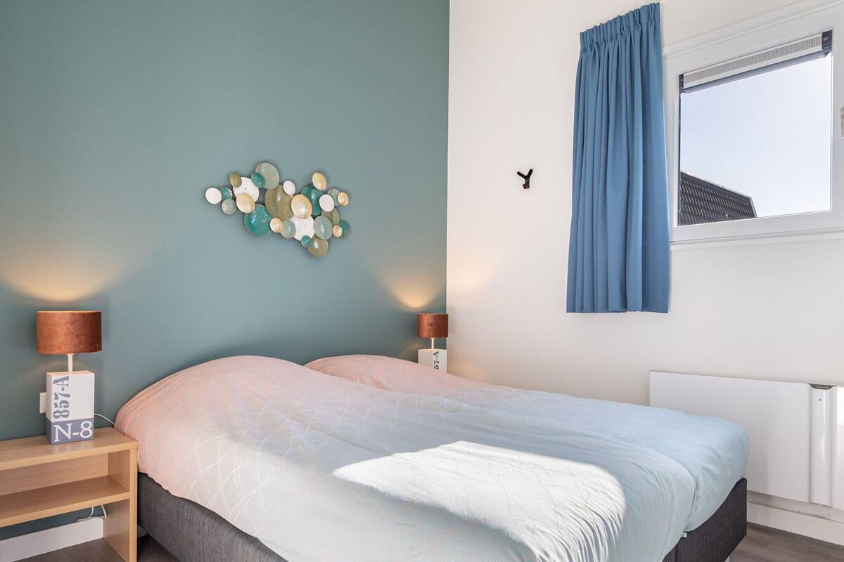 A bedroom with a bed, two bedside tables, a wall art piece, a window with curtains, and a radiator.