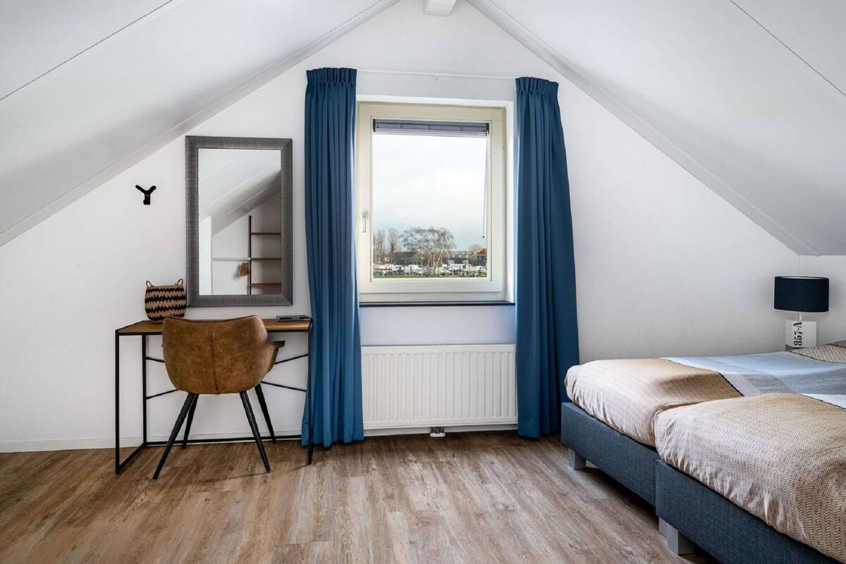 A bedroom with a sloped ceiling, a desk with a chair, a bed, a window with curtains, and a radiator.