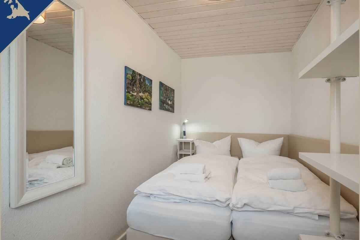 Ein Hotelzimmer mit einem Bett, einem Nachttisch mit Lampe, zwei an der Wand befestigten Bildern und einem Spiegel, der das Bett reflektiert.