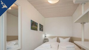 Ein Hotelzimmer mit einem Bett, einem Nachttisch mit Lampe, zwei an der Wand befestigten Bildern und einem Spiegel, der das Bett reflektiert.