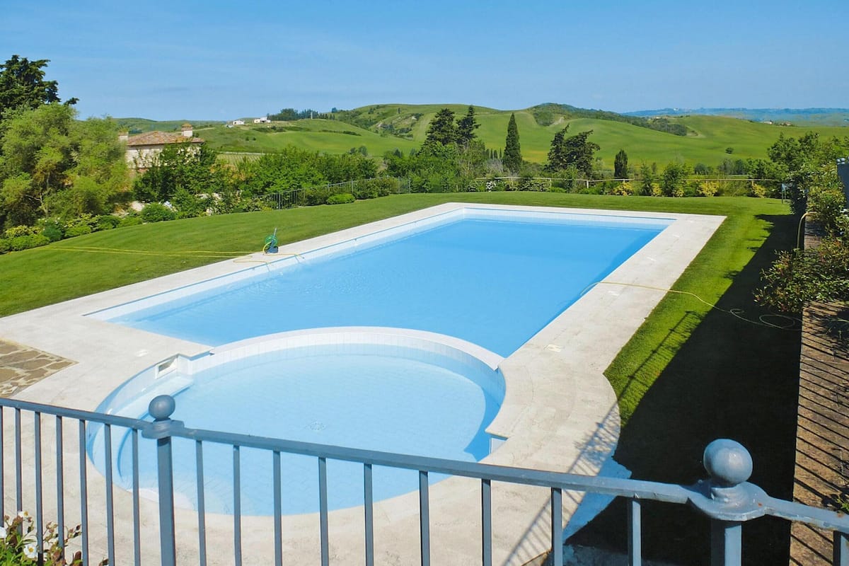 A rectangular swimming pool with a surrounding concrete deck, set in a lush green landscape with rolling hills in the background.
