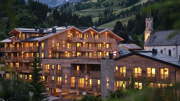 A multi-story wooden building with balconies, a church, and a mountainous backdrop. - Apartment Comfort Alpine (Muehlbach Am Hochkoenig)