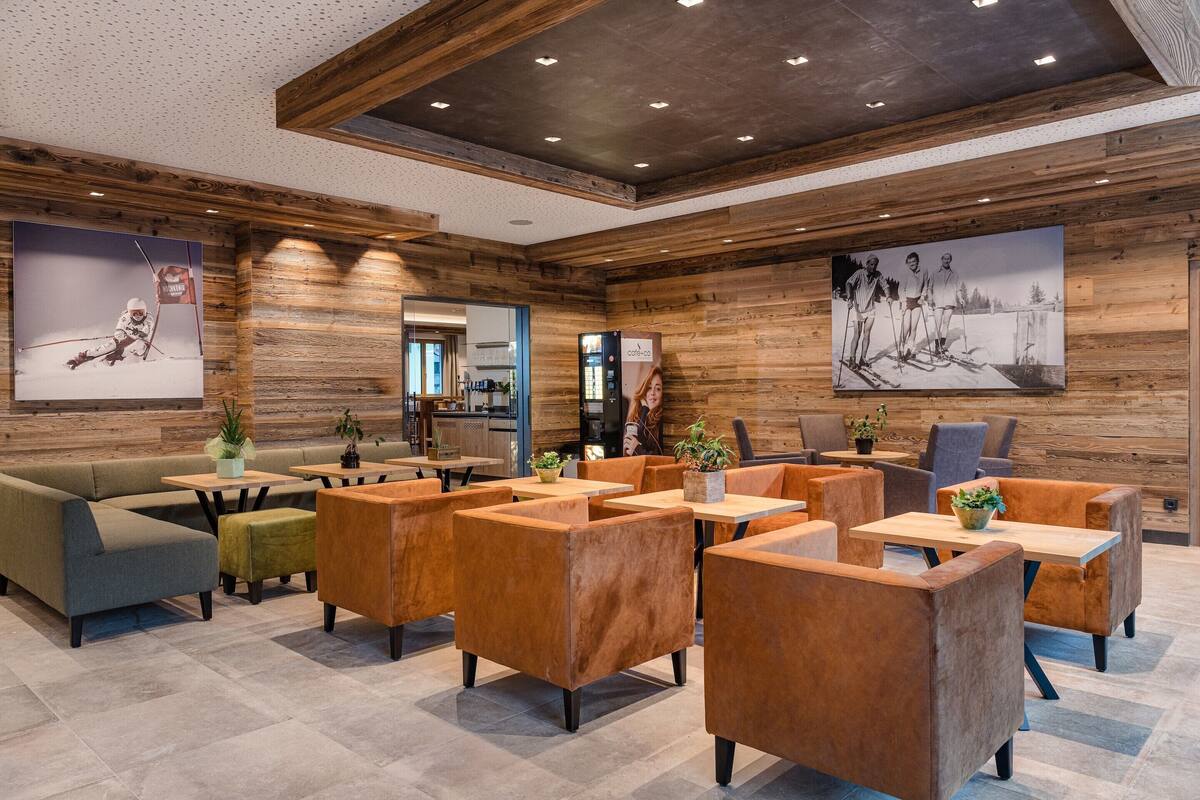 Un espacio lounge moderno con paredes de madera, diversas opciones de asientos que incluyen sofás y sillones, y una cocina al fondo.