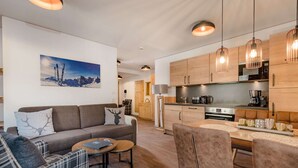 A modern living room with a sofa, a coffee table, and a dining area with a table and chairs. There is a kitchen with wooden cabinets and a stove. - Apartment Comfort Alpine (Muehlbach Am Hochkoenig)