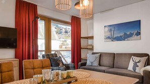 A cozy living room with a sofa, a dining table with placemats, a television, and a window with curtains. - Apartment Comfort Alpine (Muehlbach Am Hochkoenig)
