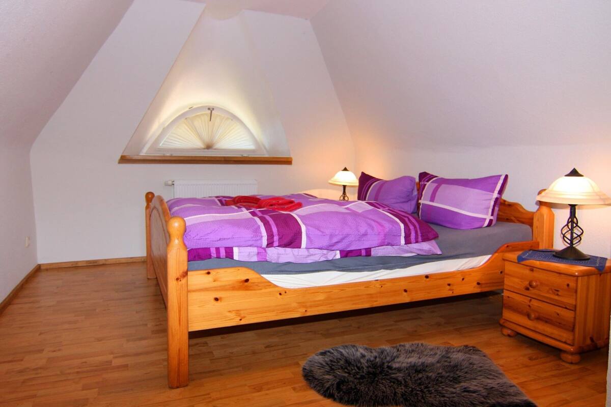A bedroom with a sloped ceiling, a wooden bed, purple bedding, two bedside lamps, a small wooden nightstand, and a fur rug.