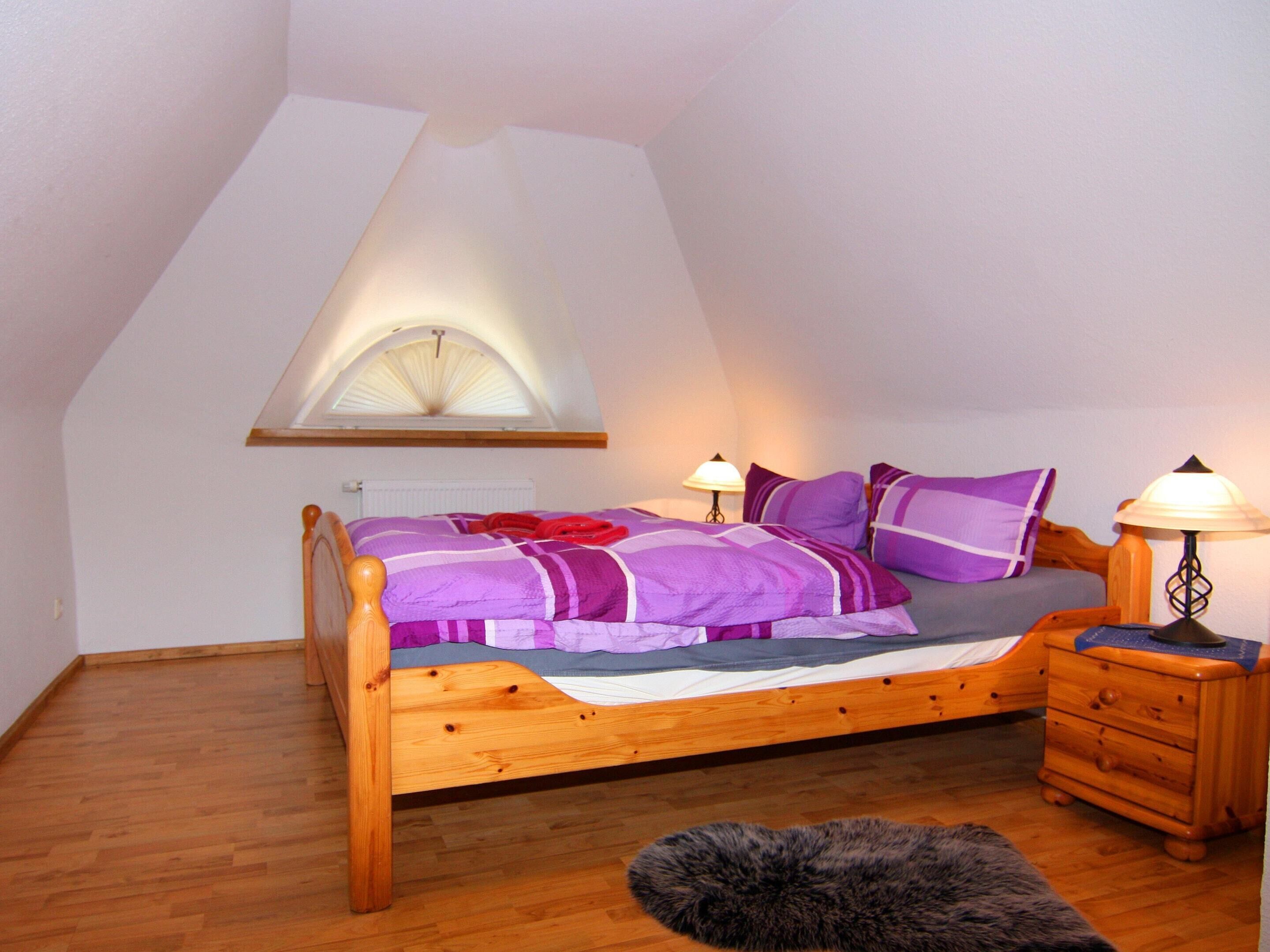 Une chambre avec un plafond en pente, un lit avec une literie violette, deux lampes de chevet, une table de chevet en bois et un tapis en fourrure.