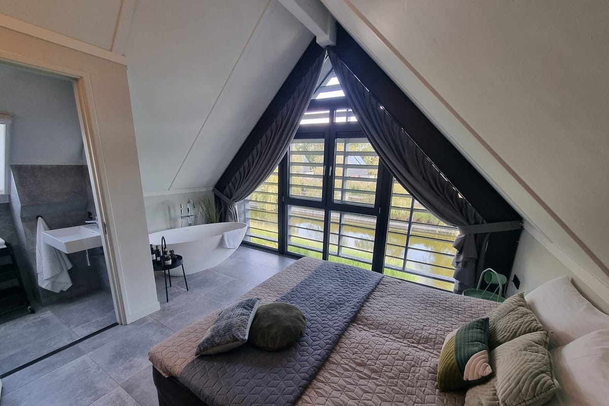 A bedroom with a large bed, a bathroom, and a view of greenery through a window.